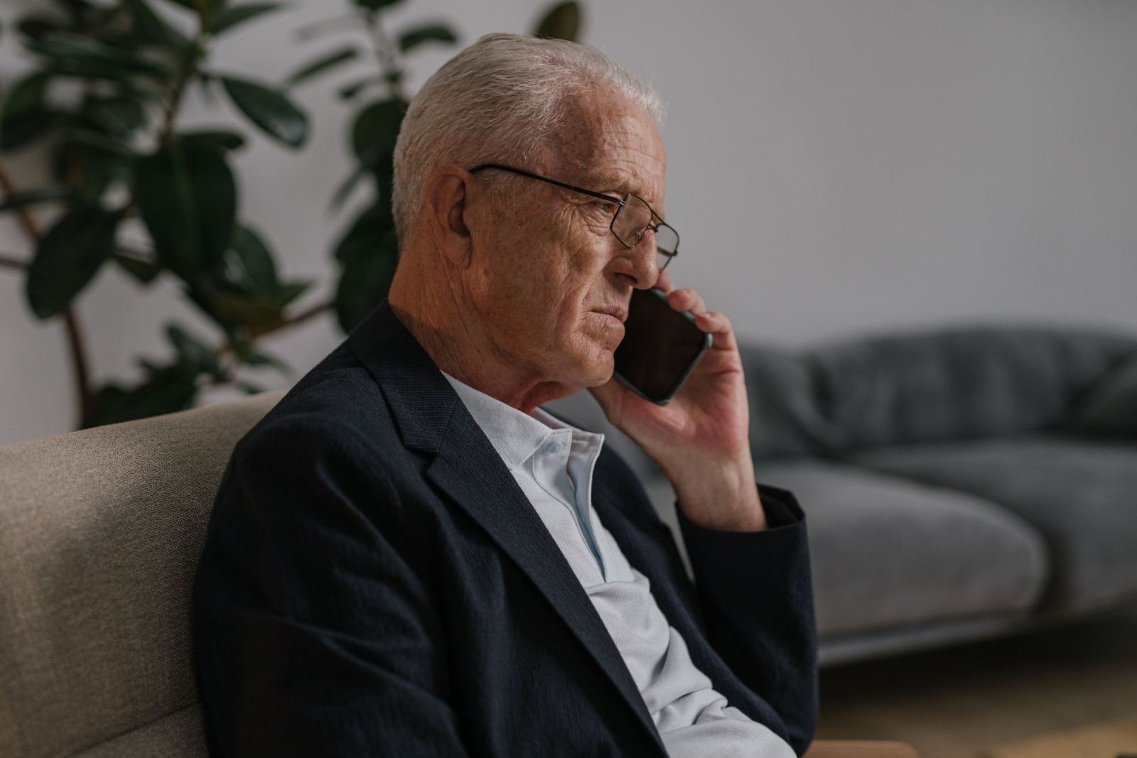 An older man in a blazer speaks on a smartphone while sitting on a couch