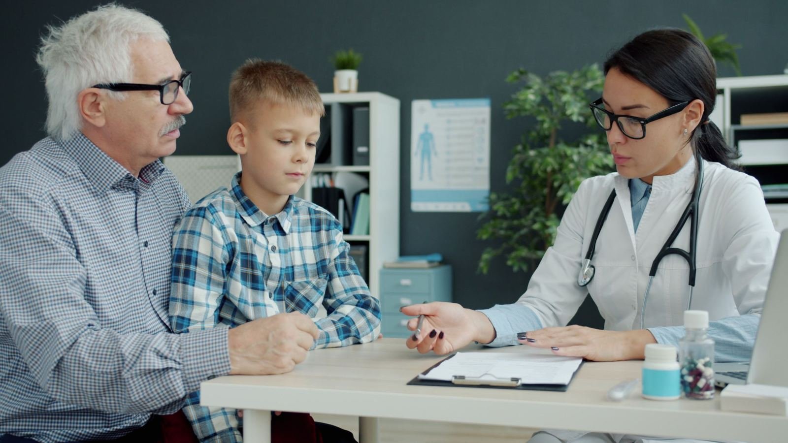 A physician in a white coat discusses paperwork with an elderly man and a young boy.