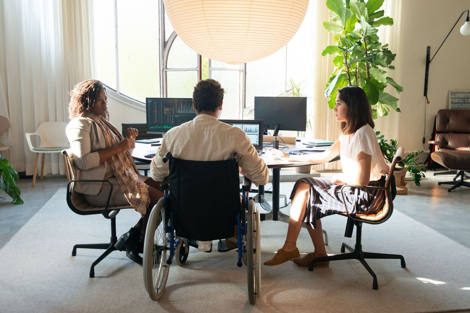 Two women and a person in a wheelchair sit at a desk with computer screens.