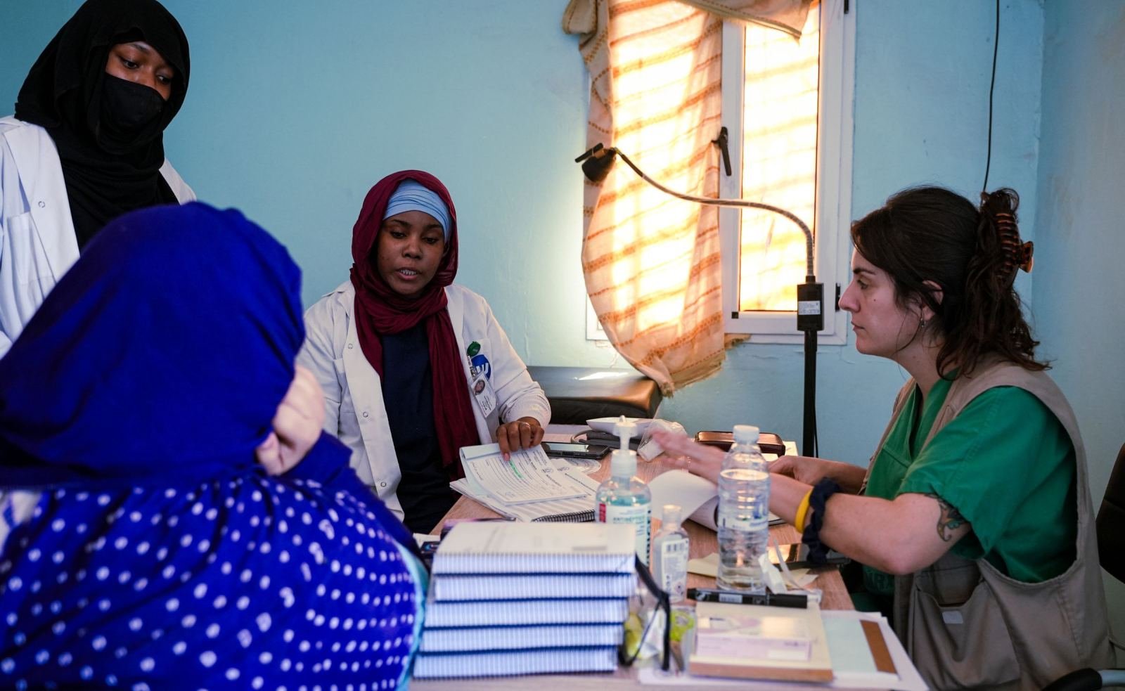 Three women in medical attire discuss paperwork at a desk in a blue room