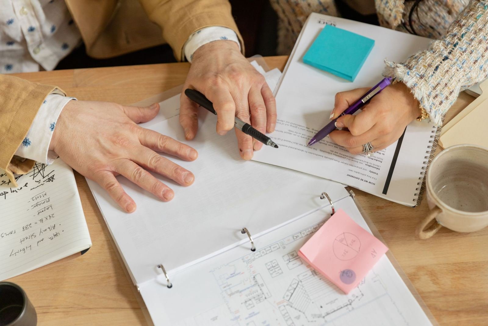 Two people use pens to point at documents and a binder on a wooden table