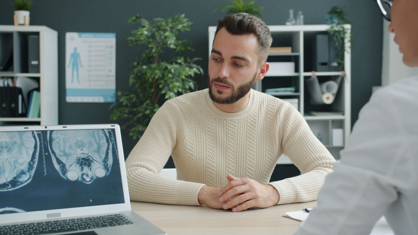A man in a beige sweater talks with a clinician in a bright office