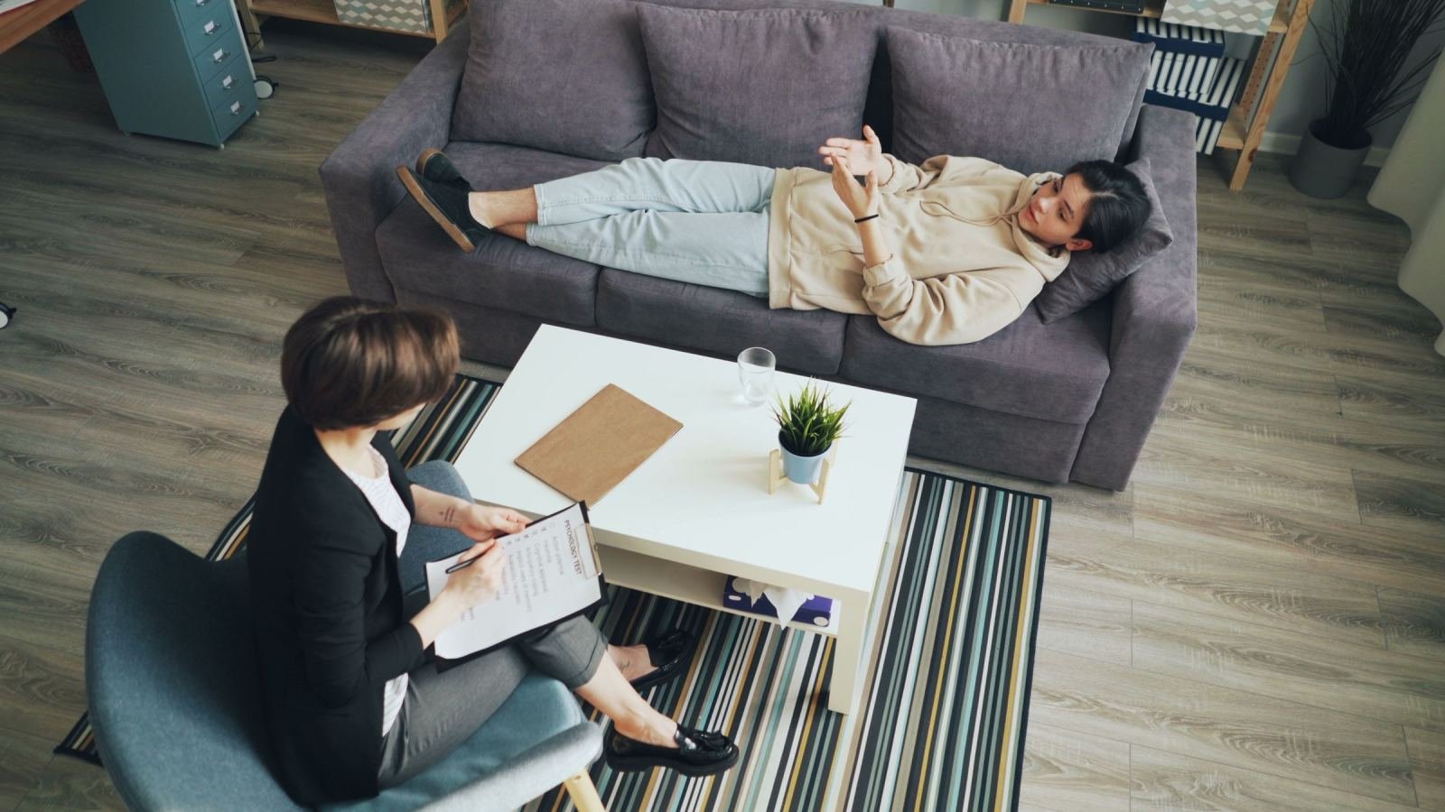 A person lies on a sofa talking to a counselor who is taking notes on a clipboard