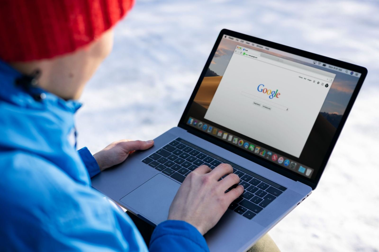 Someone wearing a red hat and blue jacket sits in the snow using a laptop.
