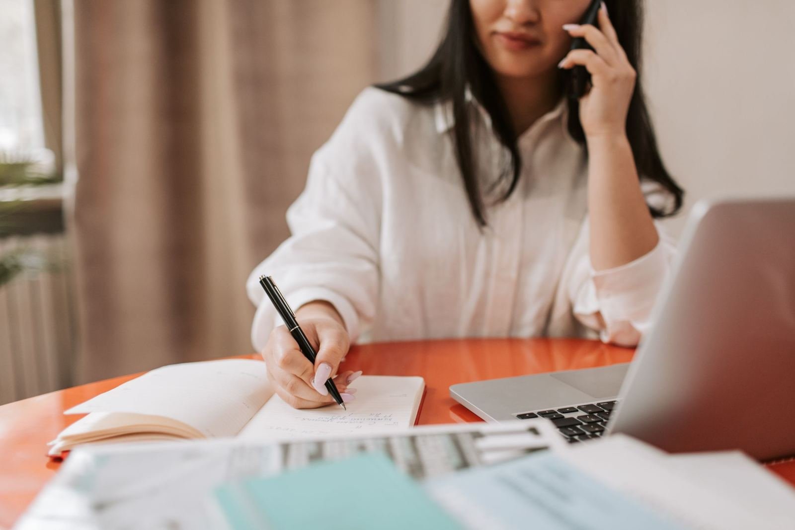 A woman on a phone call writes notes in a notebook at a table with a laptop
