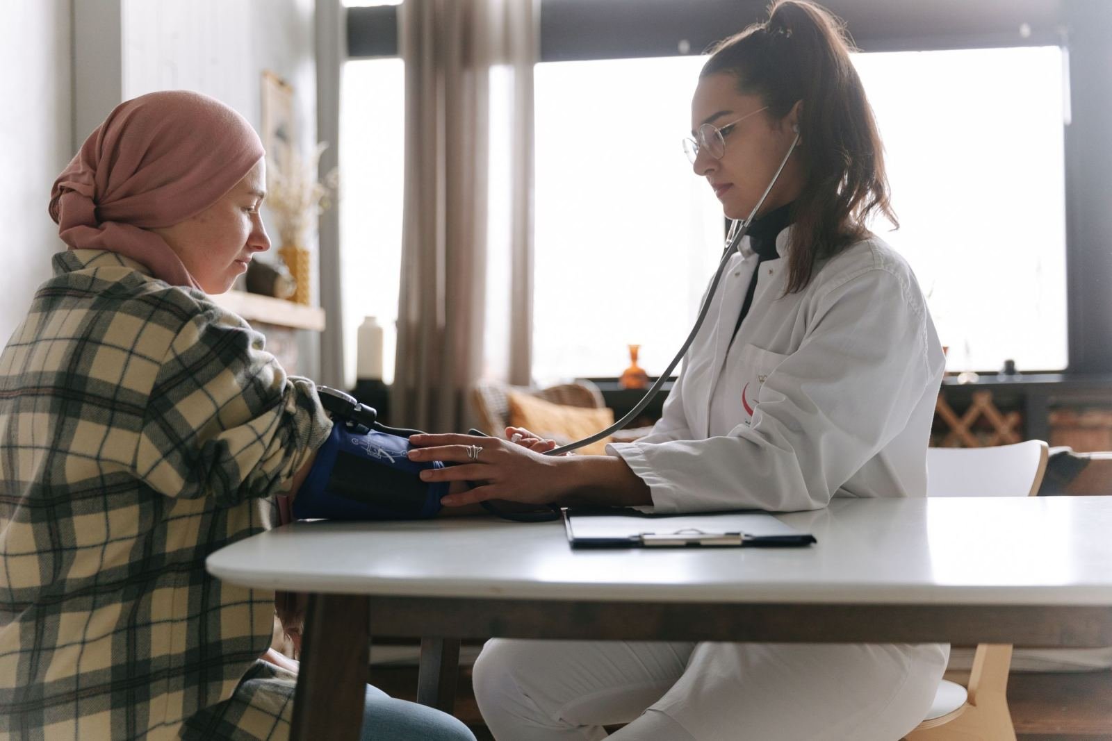 A doctor in a white coat checks the blood pressure of a woman wearing a hijab.