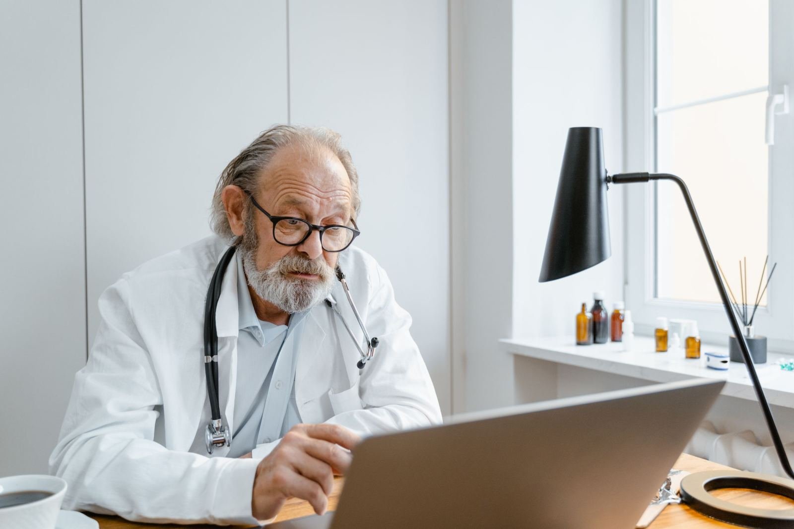 A doctor in a white coat and stethoscope looks at a laptop screen