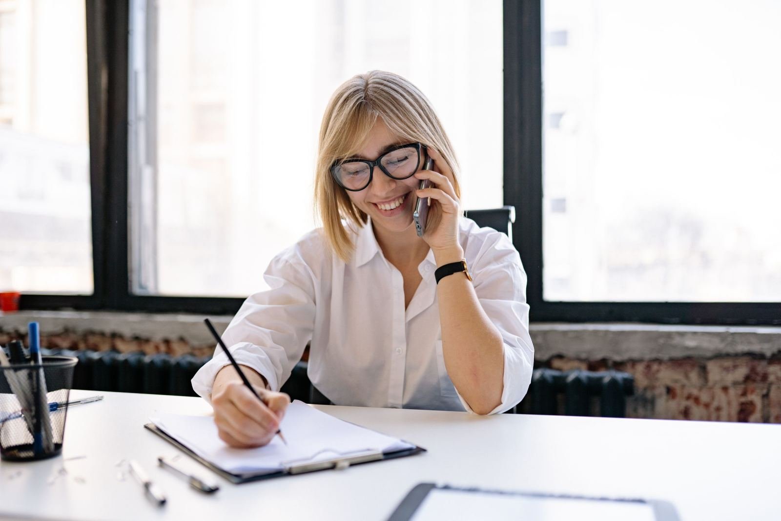 A woman with glasses smiles while talking on the phone and writing on a clipboard