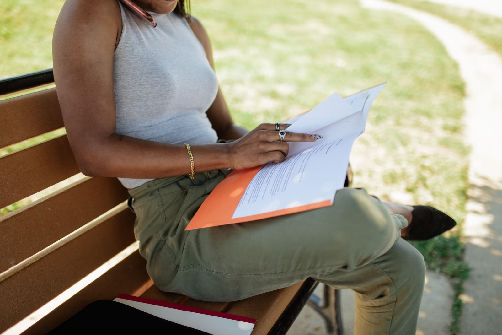 A person sitting on a park bench reading a printed document with an orange cover