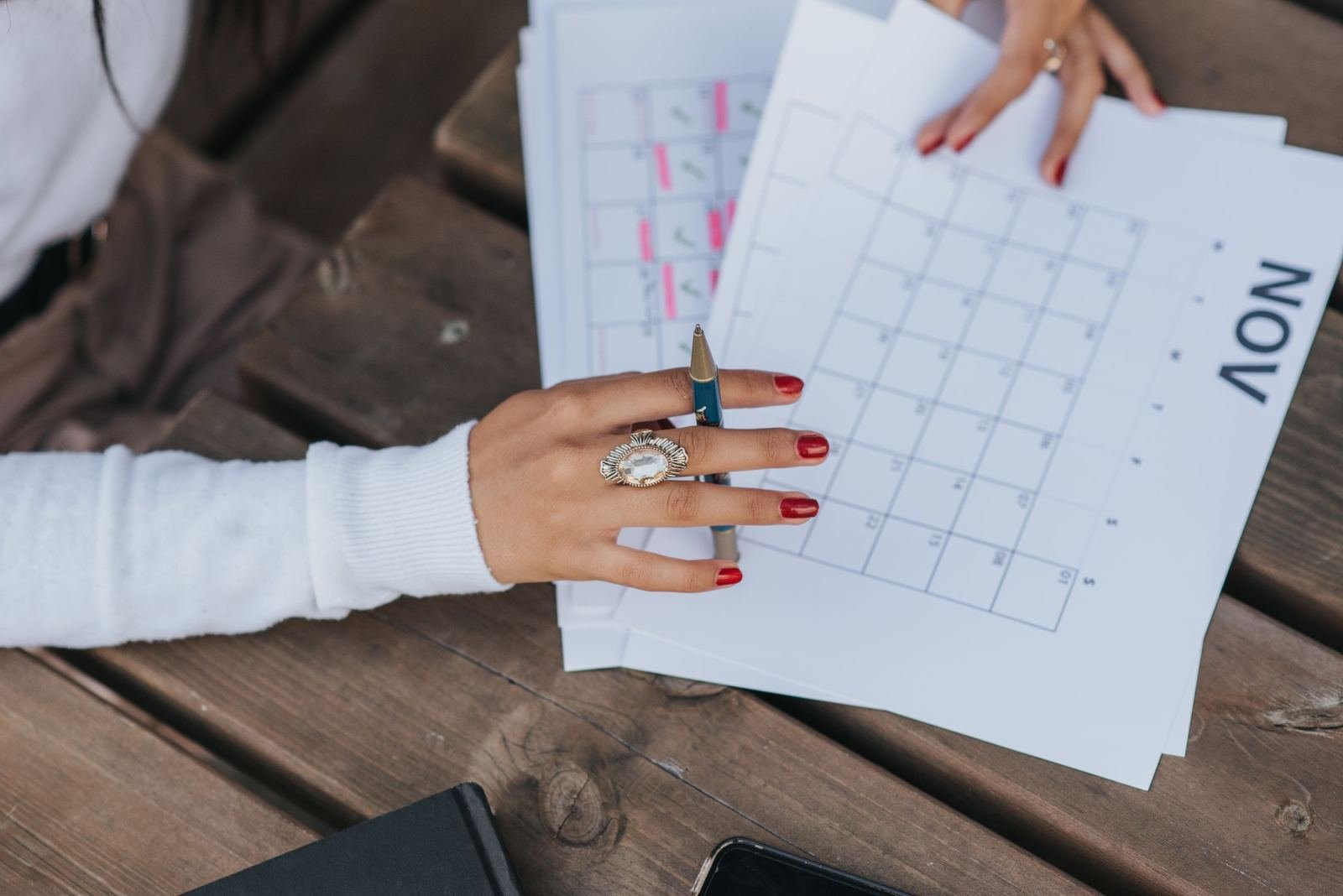 A person with a pen points to a printed calendar on a wooden table