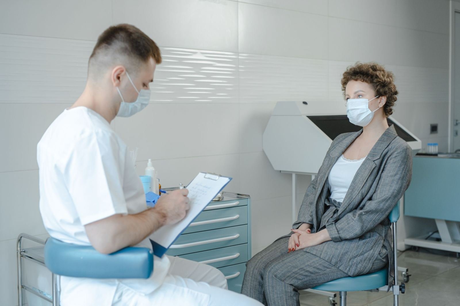 A medical professional in a mask talks with a patient in a grey suit in a clinic
