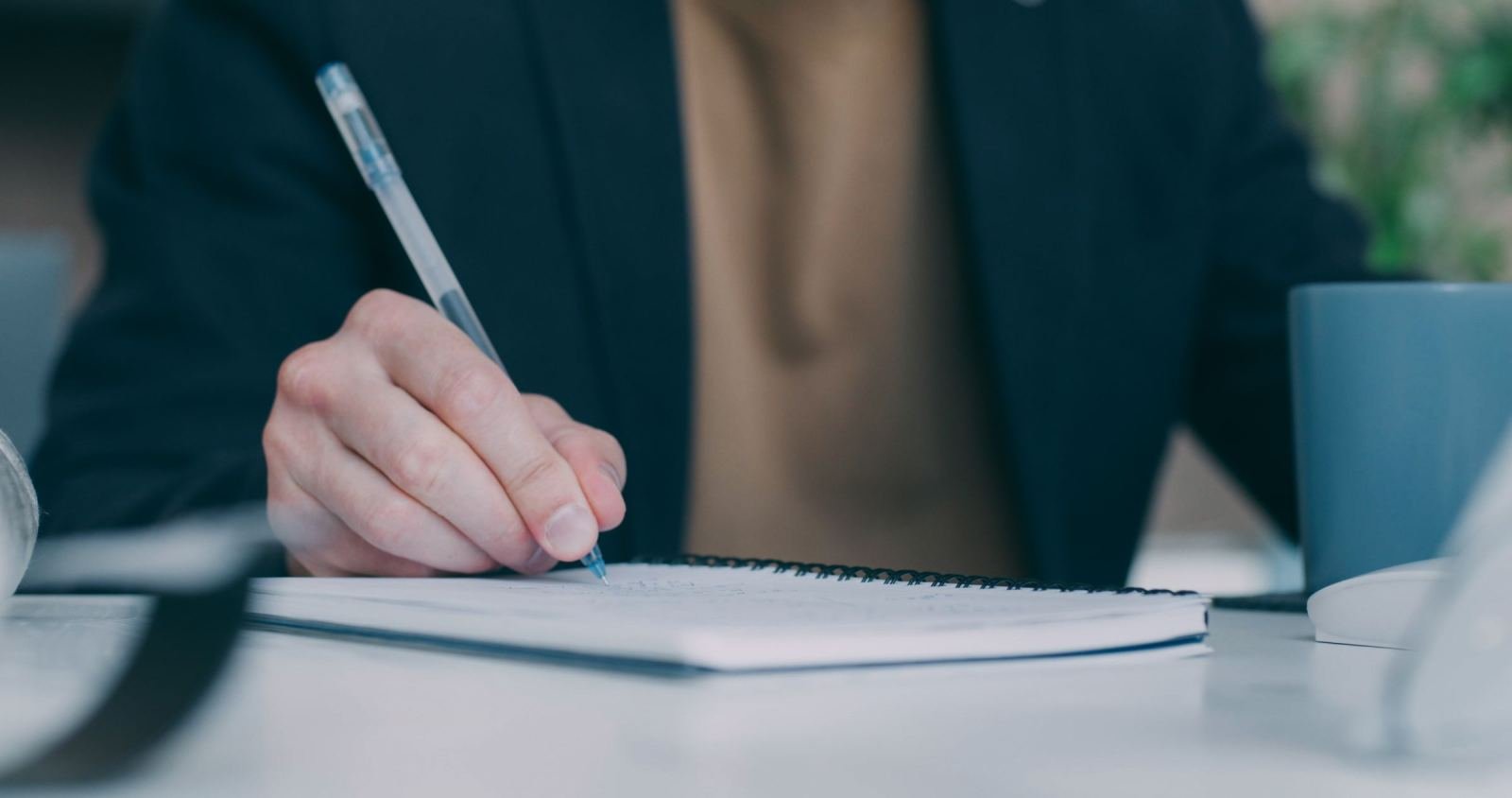 A person in a blazer writes in a spiral notebook with a blue pen