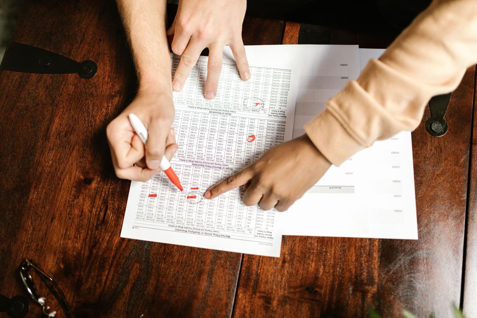 Two people use a red marker to review a budget spreadsheet on a wooden table