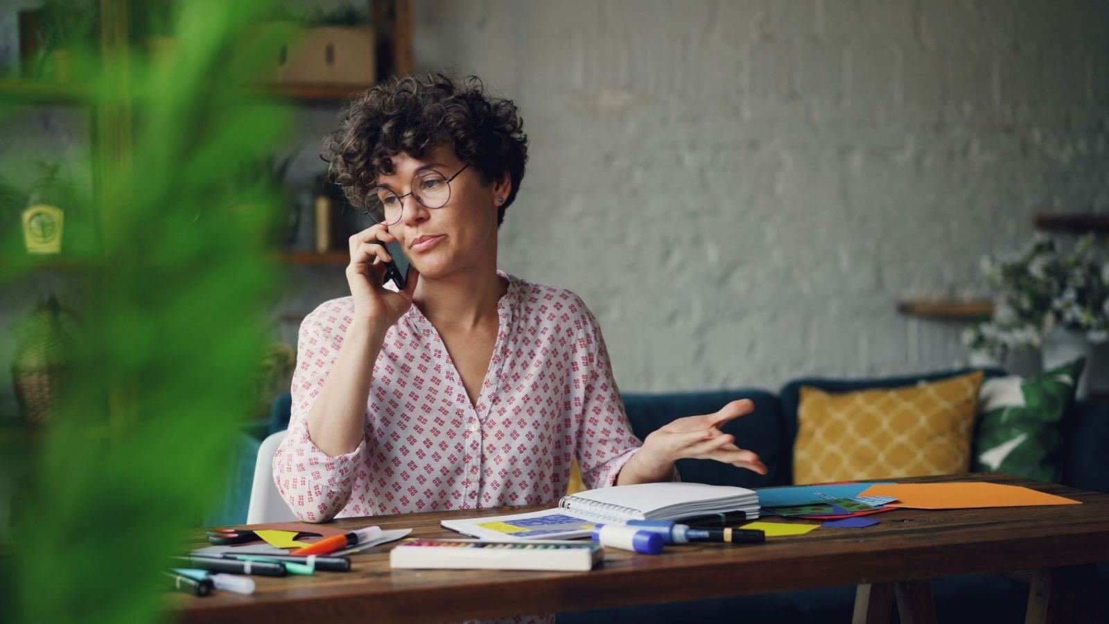 A woman with glasses talks on a phone while sitting at a desk with a notebook and markers