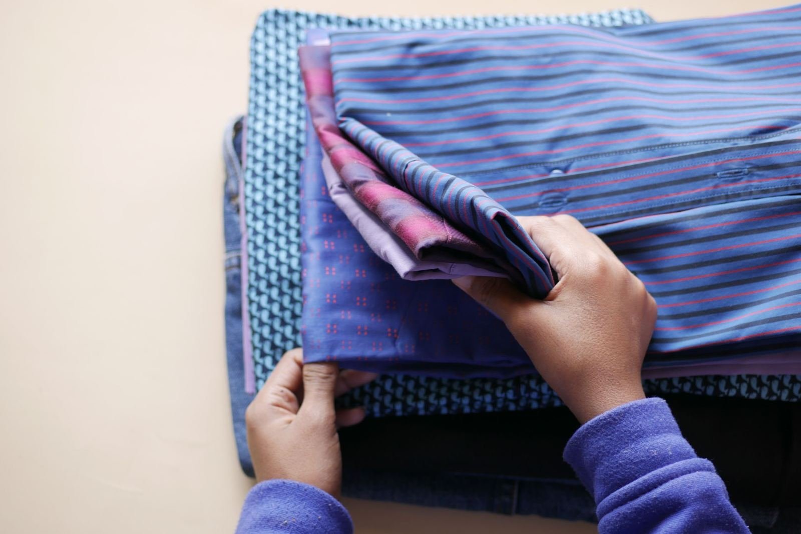 Hands folding a stack of blue and purple clothing on a light surface
