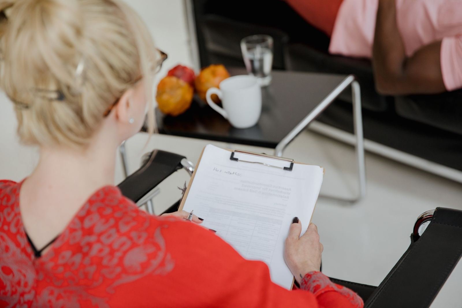 A woman in a red top holds a clipboard while sitting across from another person in an office