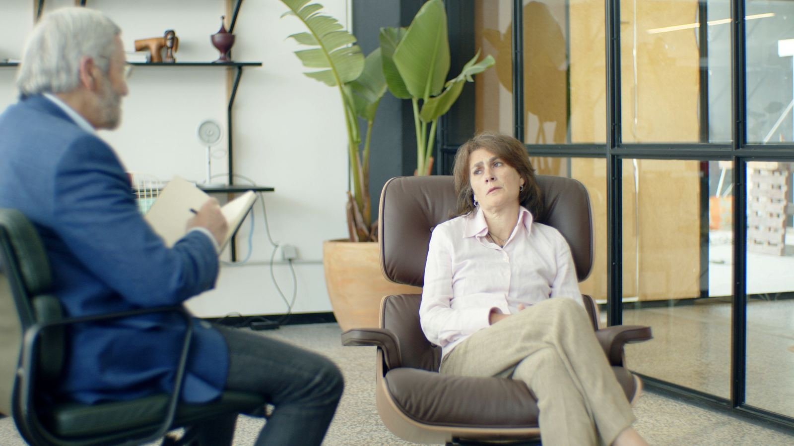 A therapist with a notebook talks to a woman sitting in a leather armchair