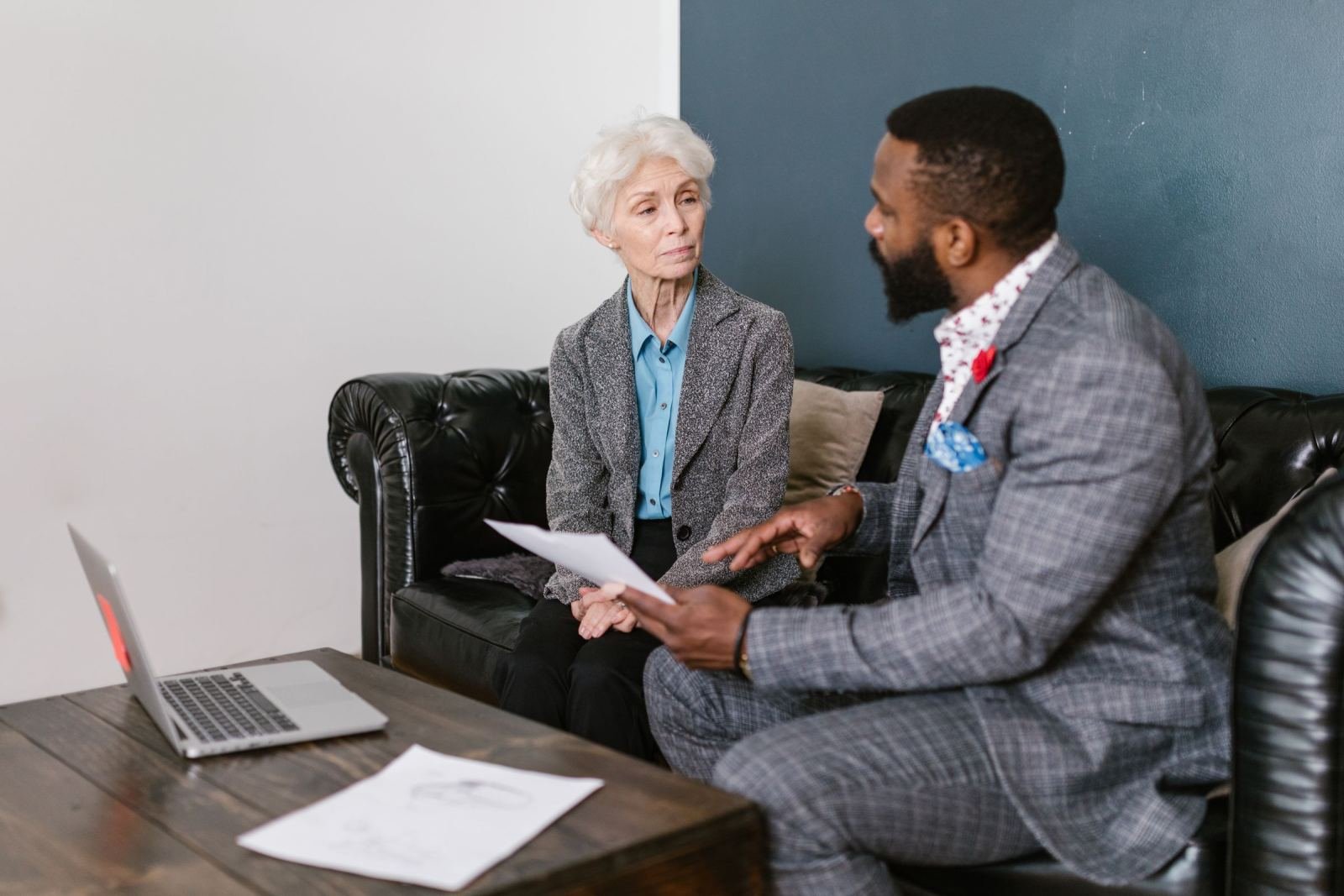 A man in a suit and a woman with white hair talk while sitting on a black leather sofa