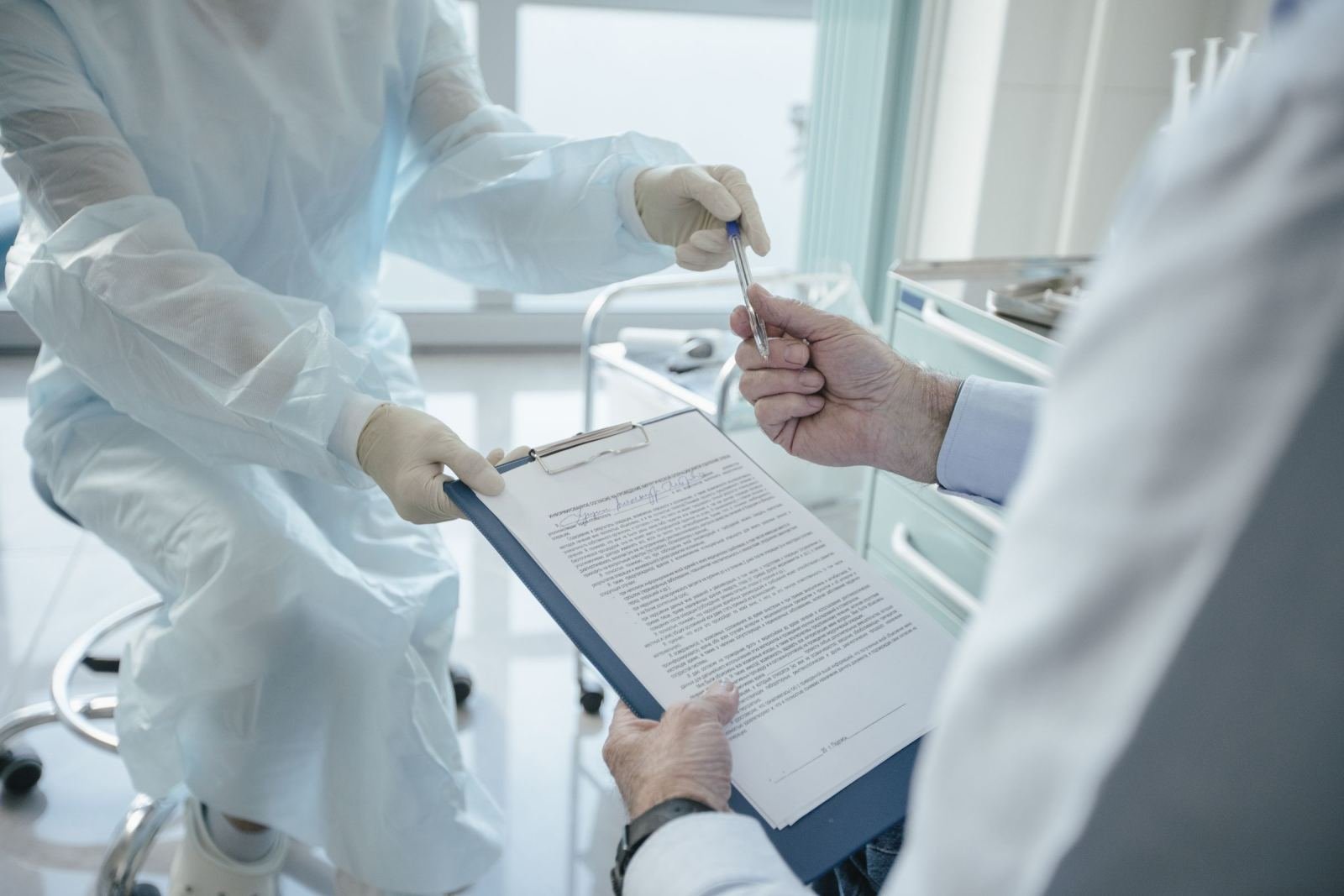 A medical professional in a blue gown hands a pen to someone holding a clipboard