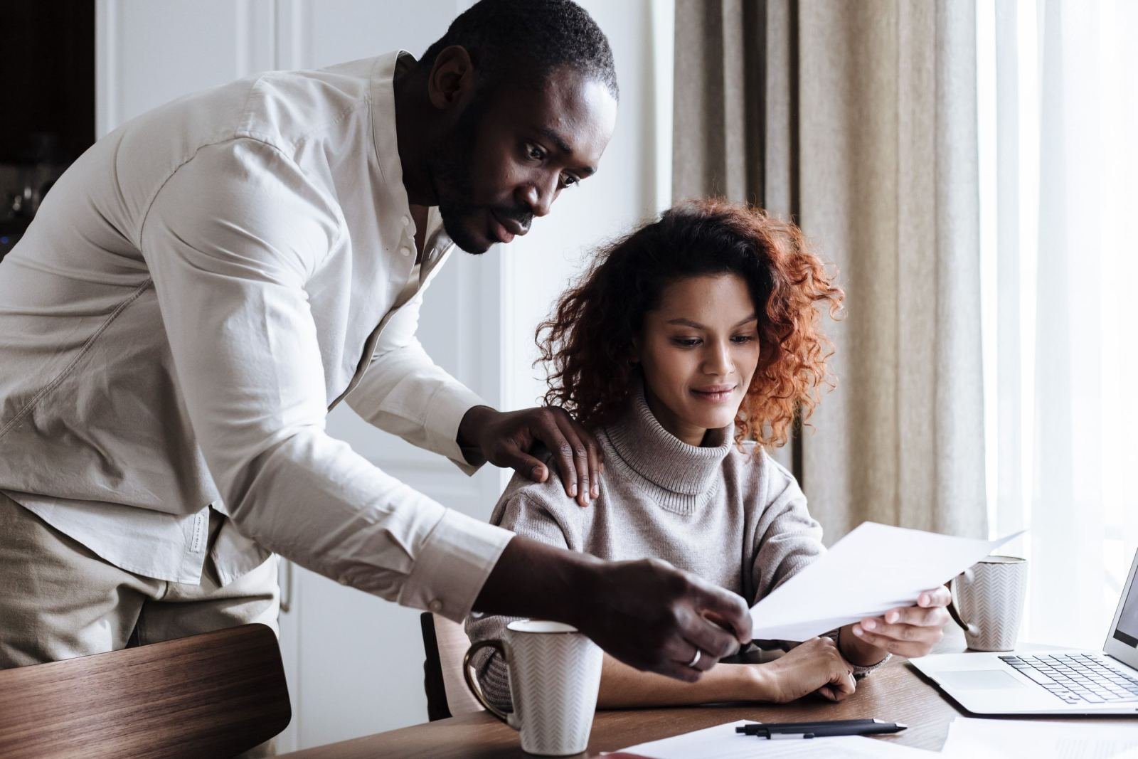 A couple sits at a desk and looks over a paper while using a laptop.