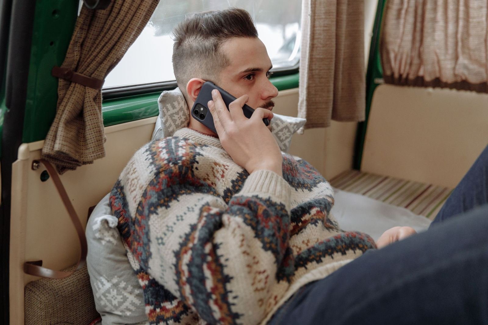 A man in a patterned sweater talks on a phone while sitting in a vehicle