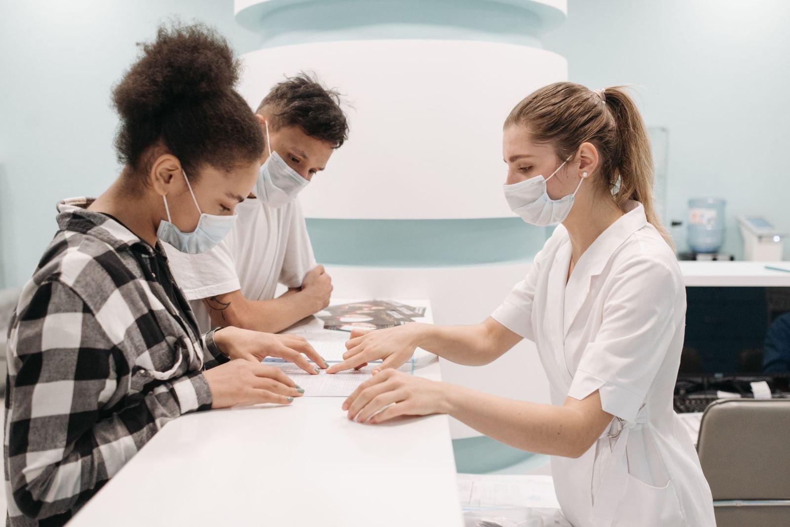 A masked clinician in a white coat looks at papers with two people at a counter.