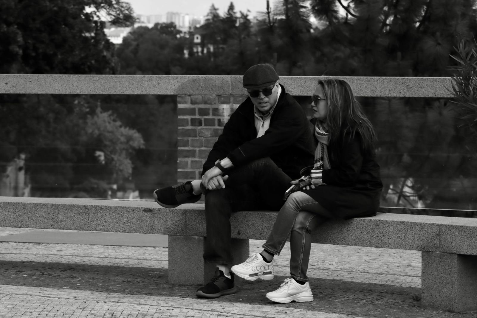 A man and woman wearing sunglasses sit and talk on a stone park bench