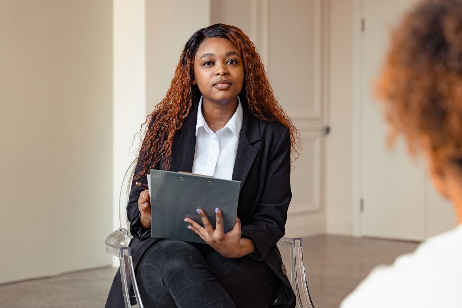 A professional woman with a clipboard sits in a clear chair during a meeting