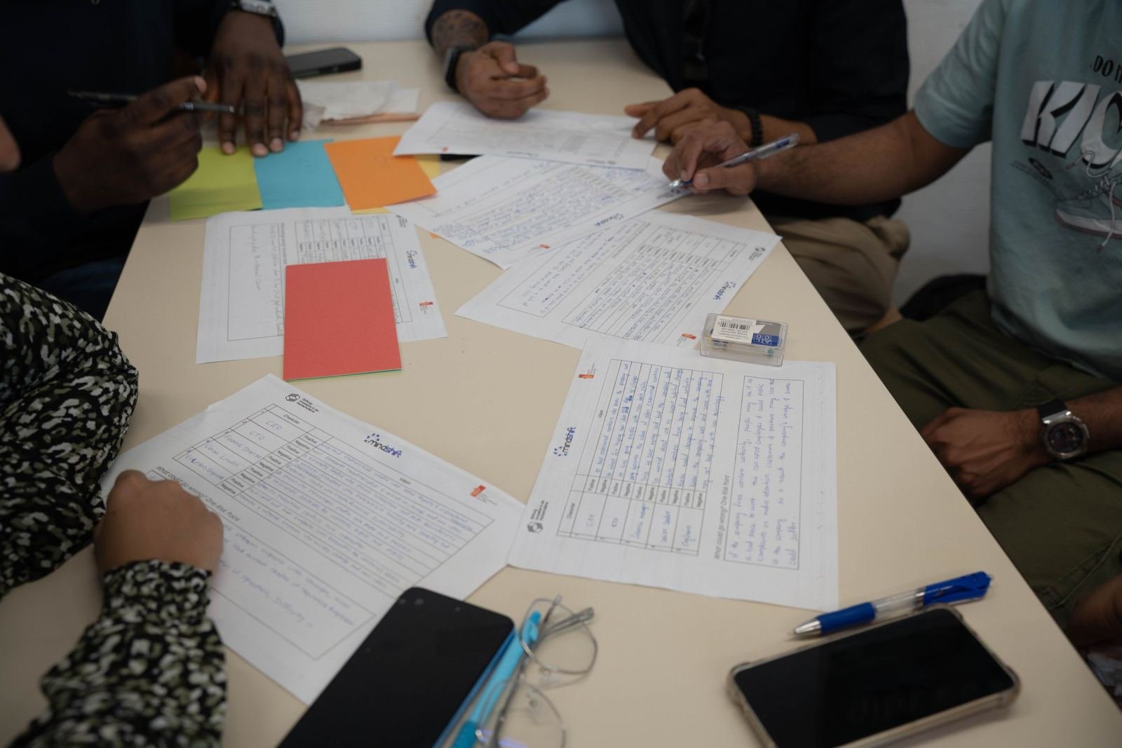 A white table holds documents and handwritten lists. Three people are reviewing them.