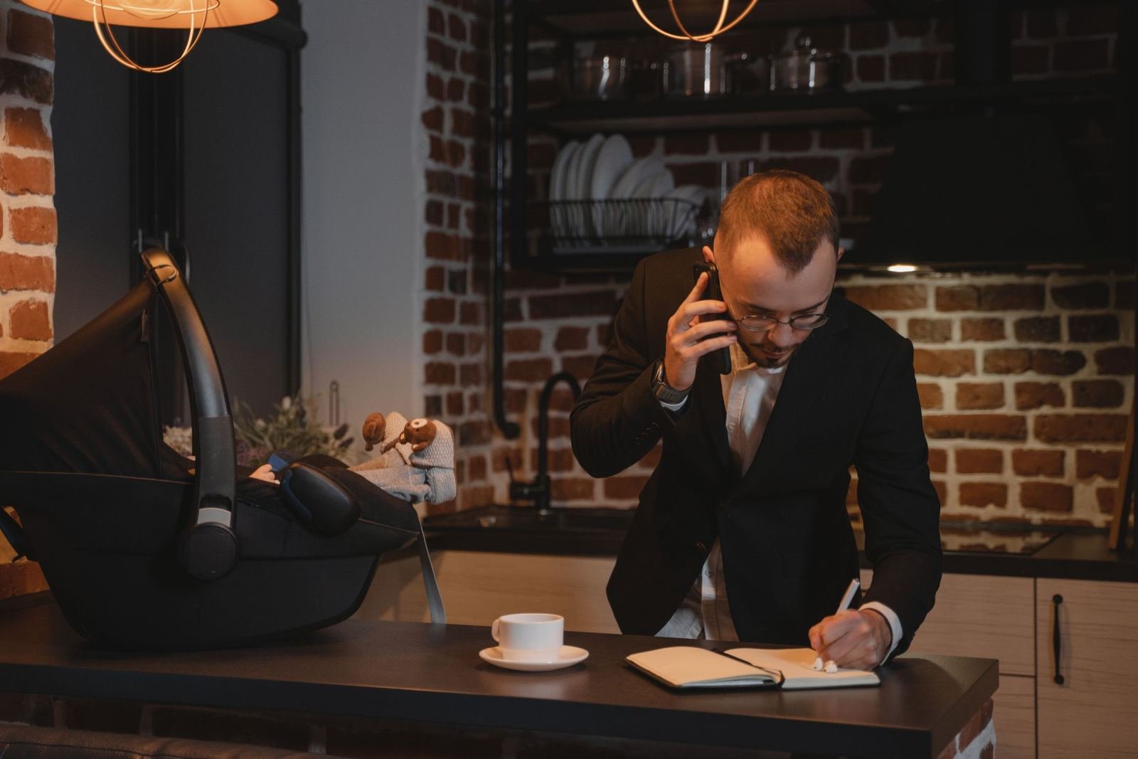 A man in a suit talks on the phone and writes in a notebook next to a baby in a car seat