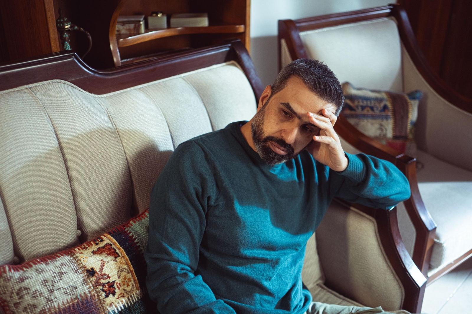 A man in a teal sweater sits on a sofa with his hand on his forehead