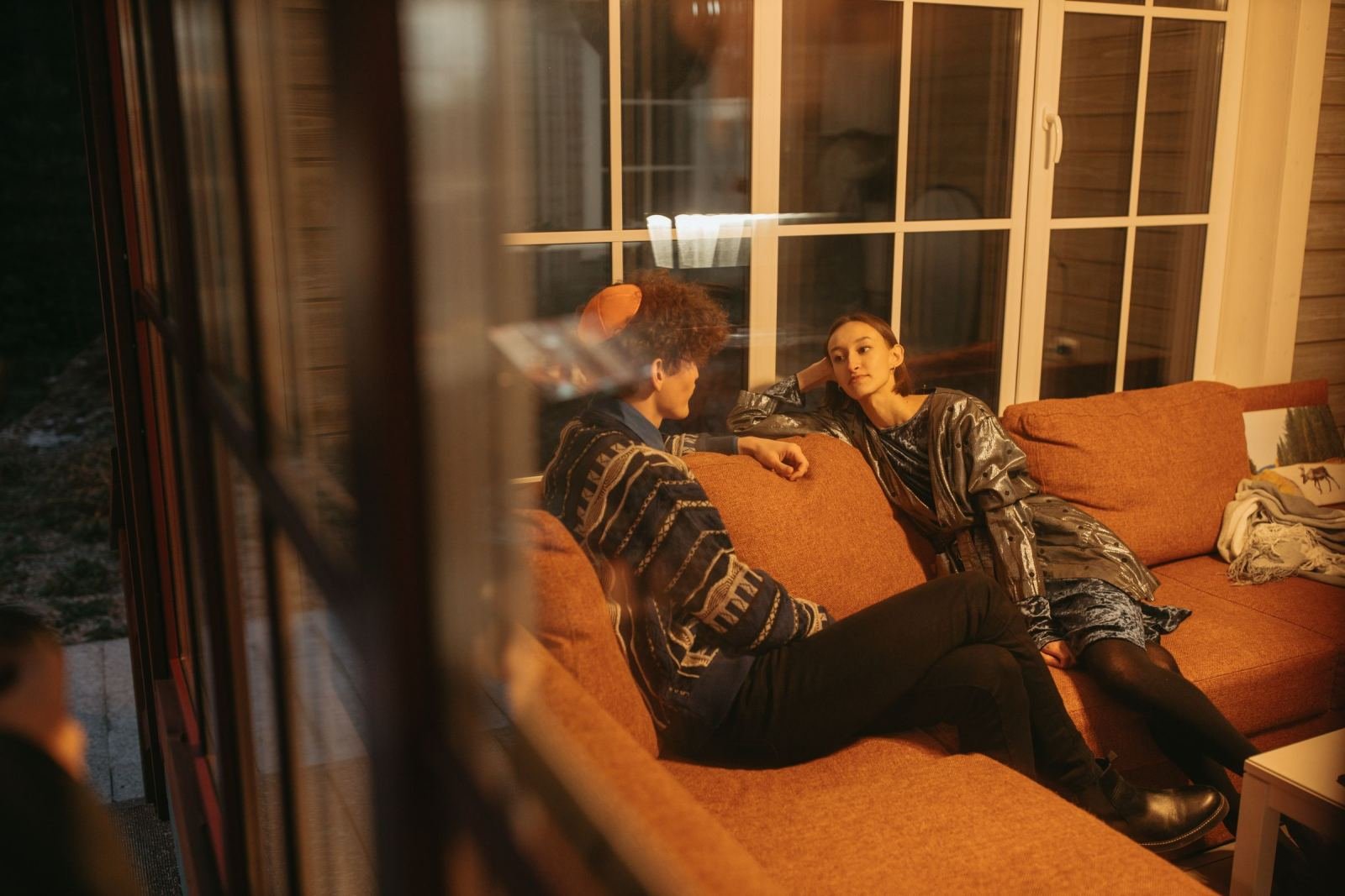 Two people talking while sitting on an orange sofa, seen through a window
