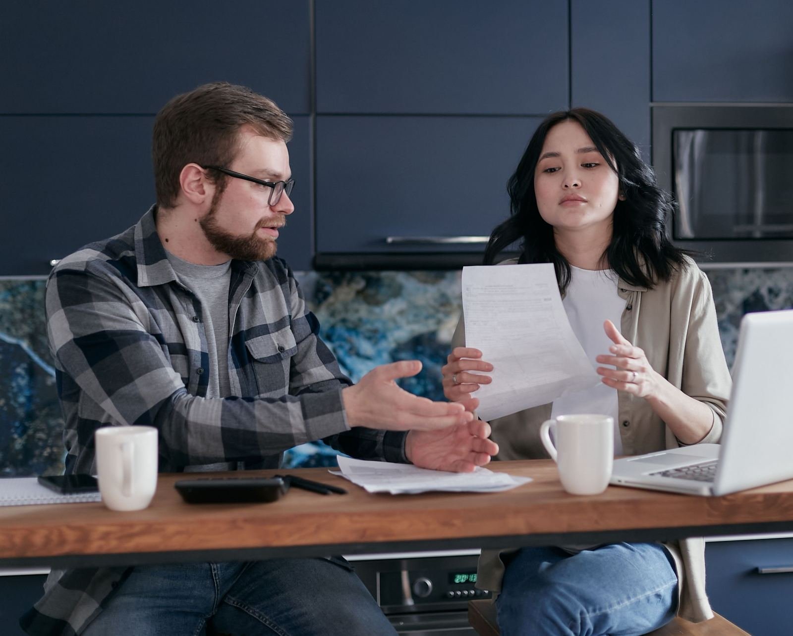 A man and woman review a document together at a kitchen table with a laptop