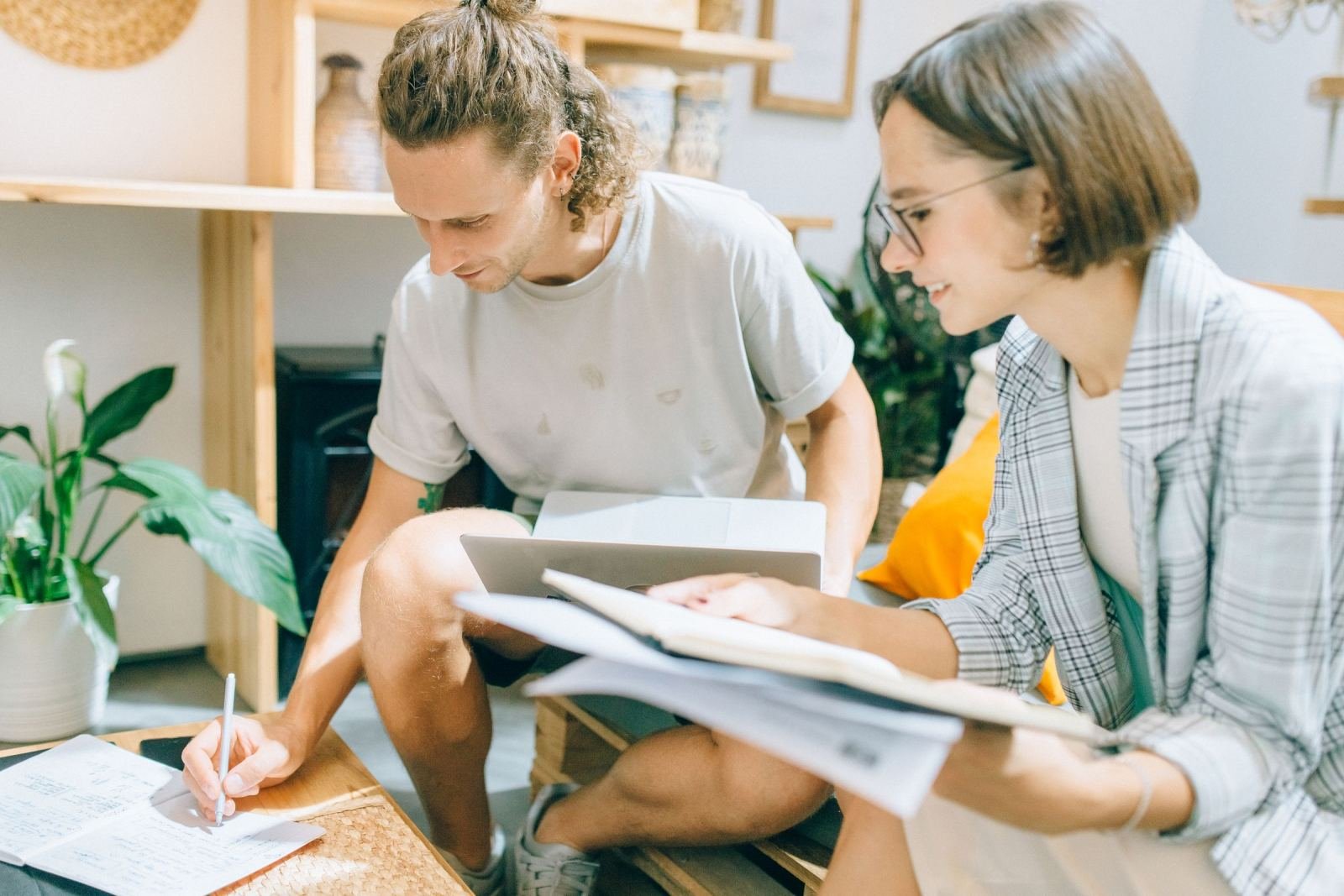 A man writes in a notebook while a woman looks on and holds papers