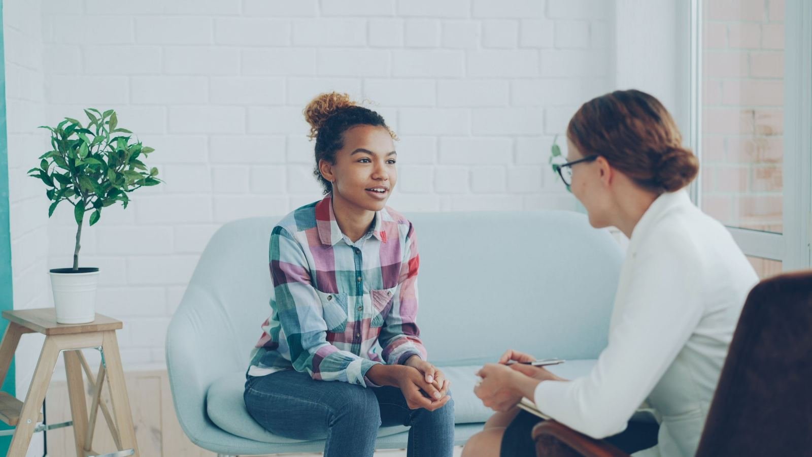 A teenage girl in a plaid shirt talks with a woman in a white blazer in a bright office