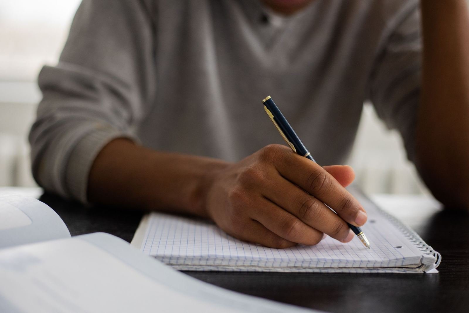 A person writes in a lined notebook with a blue pen while sitting at a desk