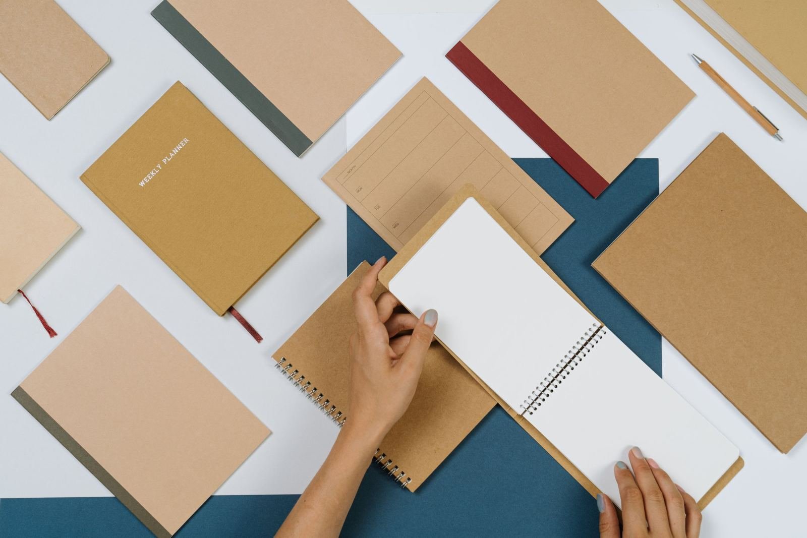 Hands opening a blank spiral notebook surrounded by other brown notebooks and a pencil