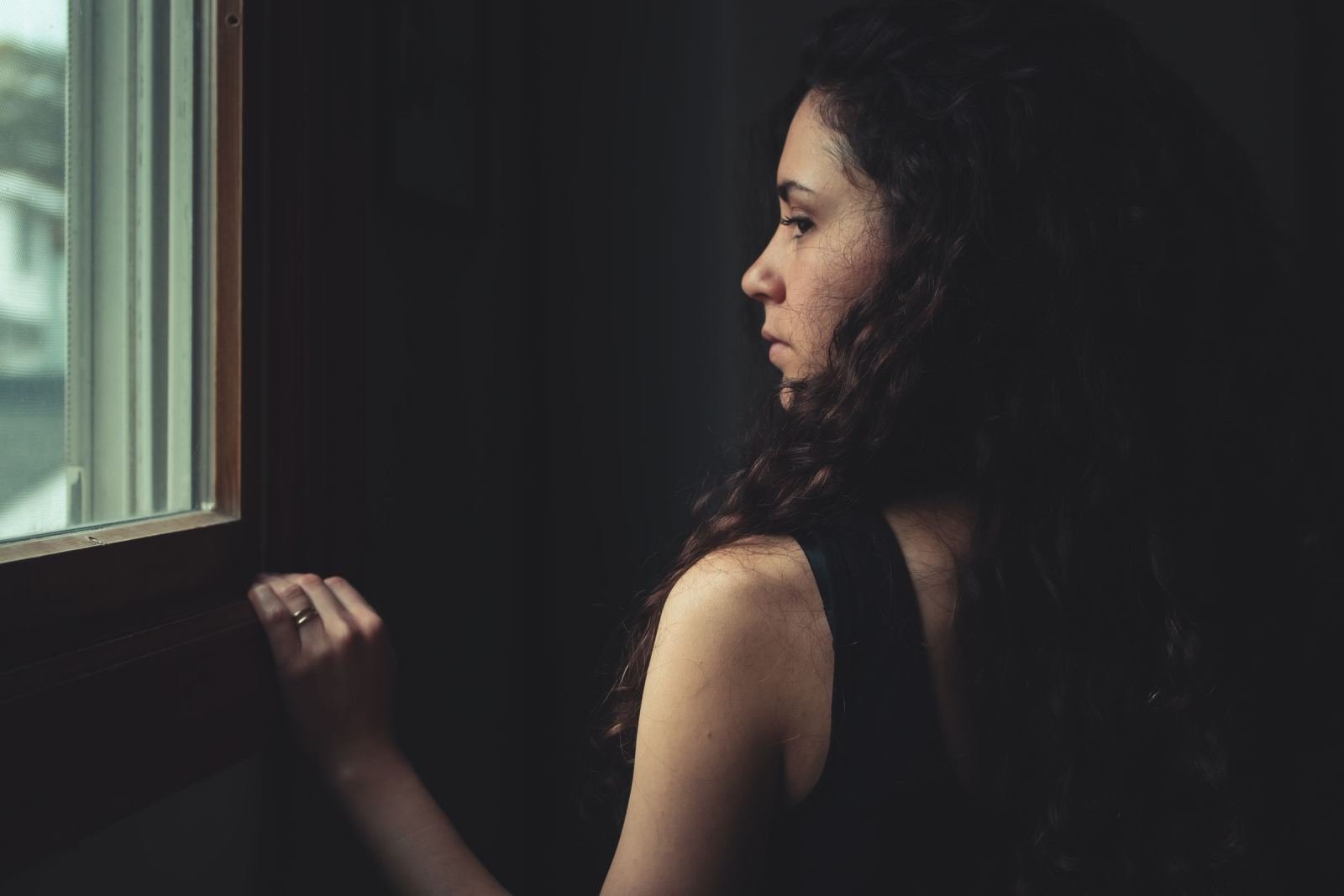 A woman with curly hair looks out a window in a dimly lit room
