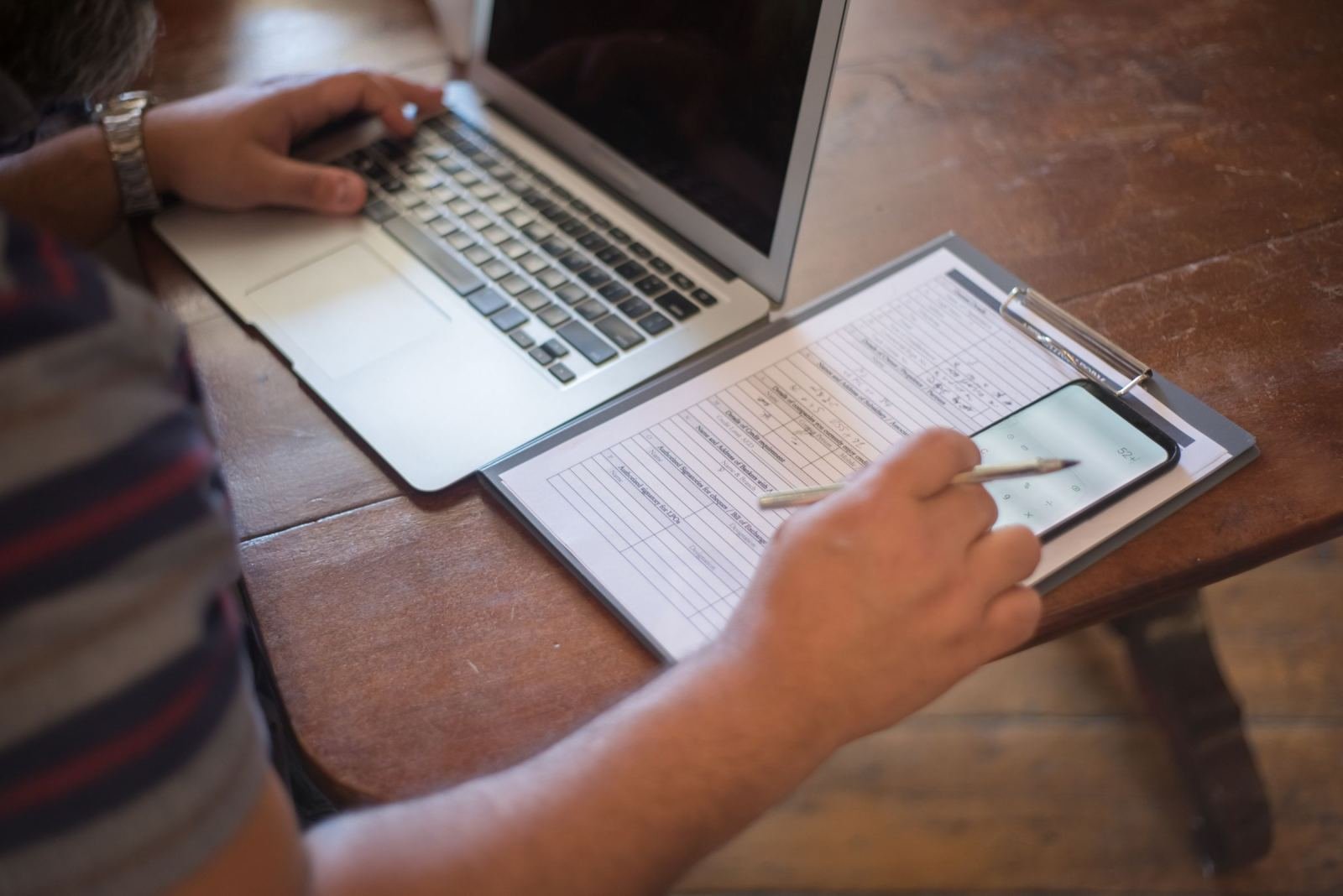 A person using a laptop and reviewing a form on a clipboard at a wooden table