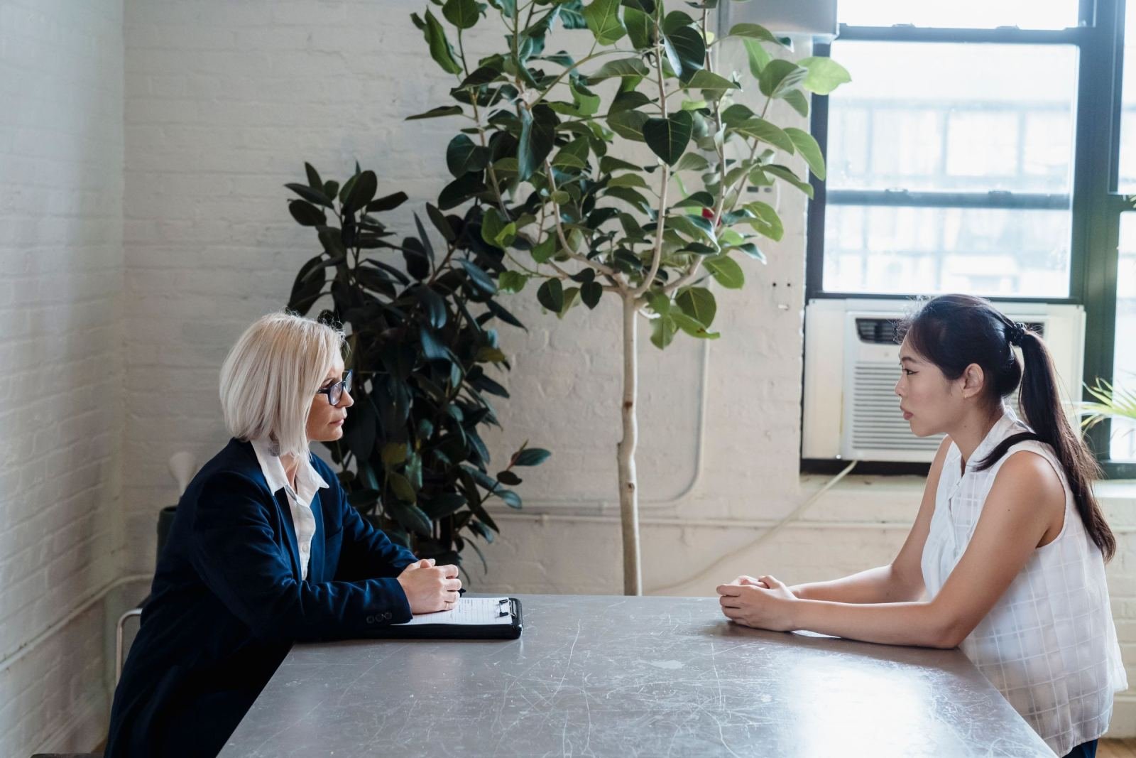 Two women sit at a table talking, one with a clipboard