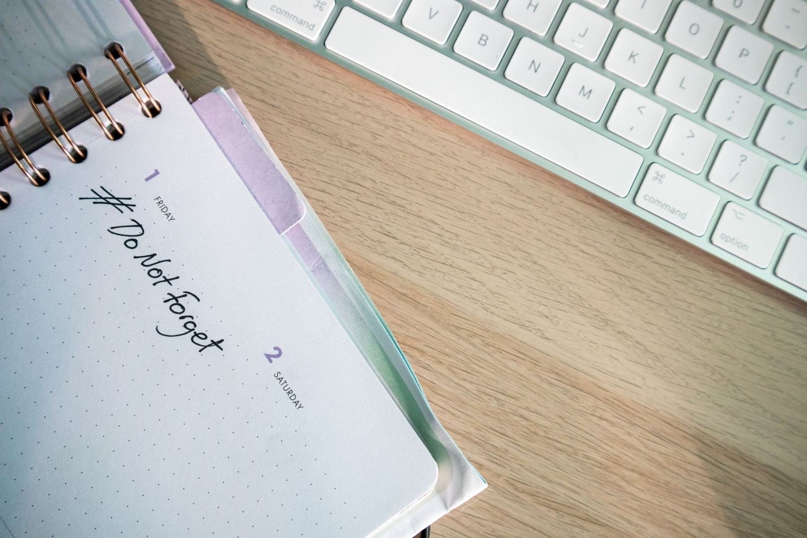 An open planner on a wooden desk next to a white keyboard with the words # Do Not Forget