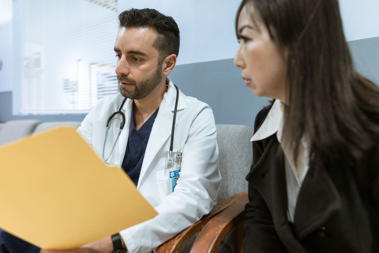 A clinician in a white coat holds a yellow folder while talking to a woman