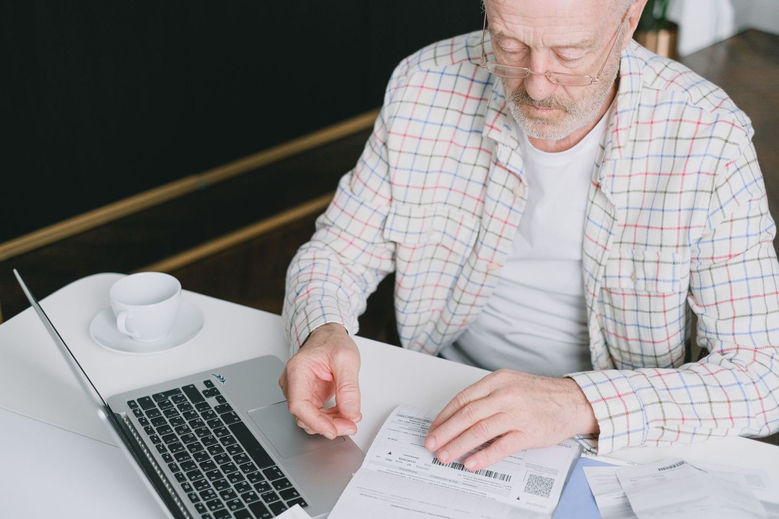 An older man in a plaid shirt reviews a document at a desk with a laptop