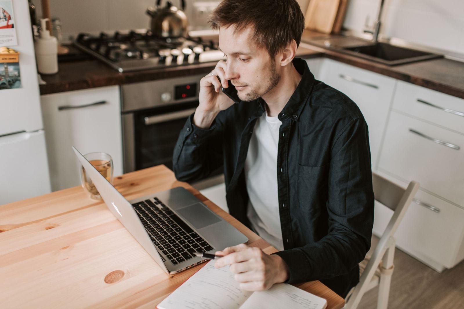 A man on a phone call writes in a notebook with a laptop open on a kitchen table