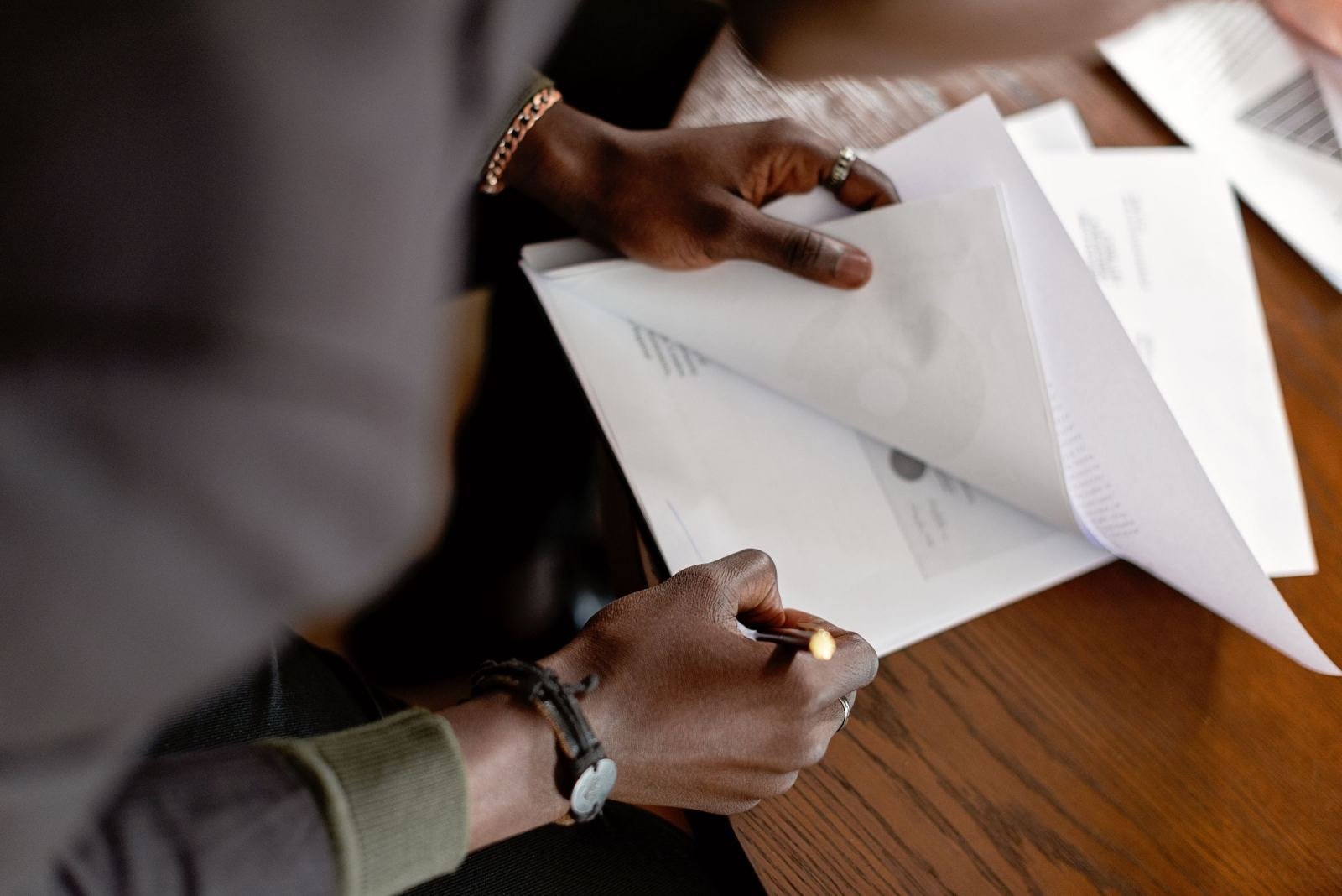 A person reviews and takes notes on printed documents at a wooden table
