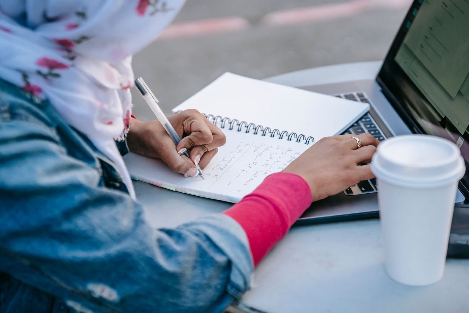 A person wearing a headscarf writes in a notebook next to a laptop and coffee cup