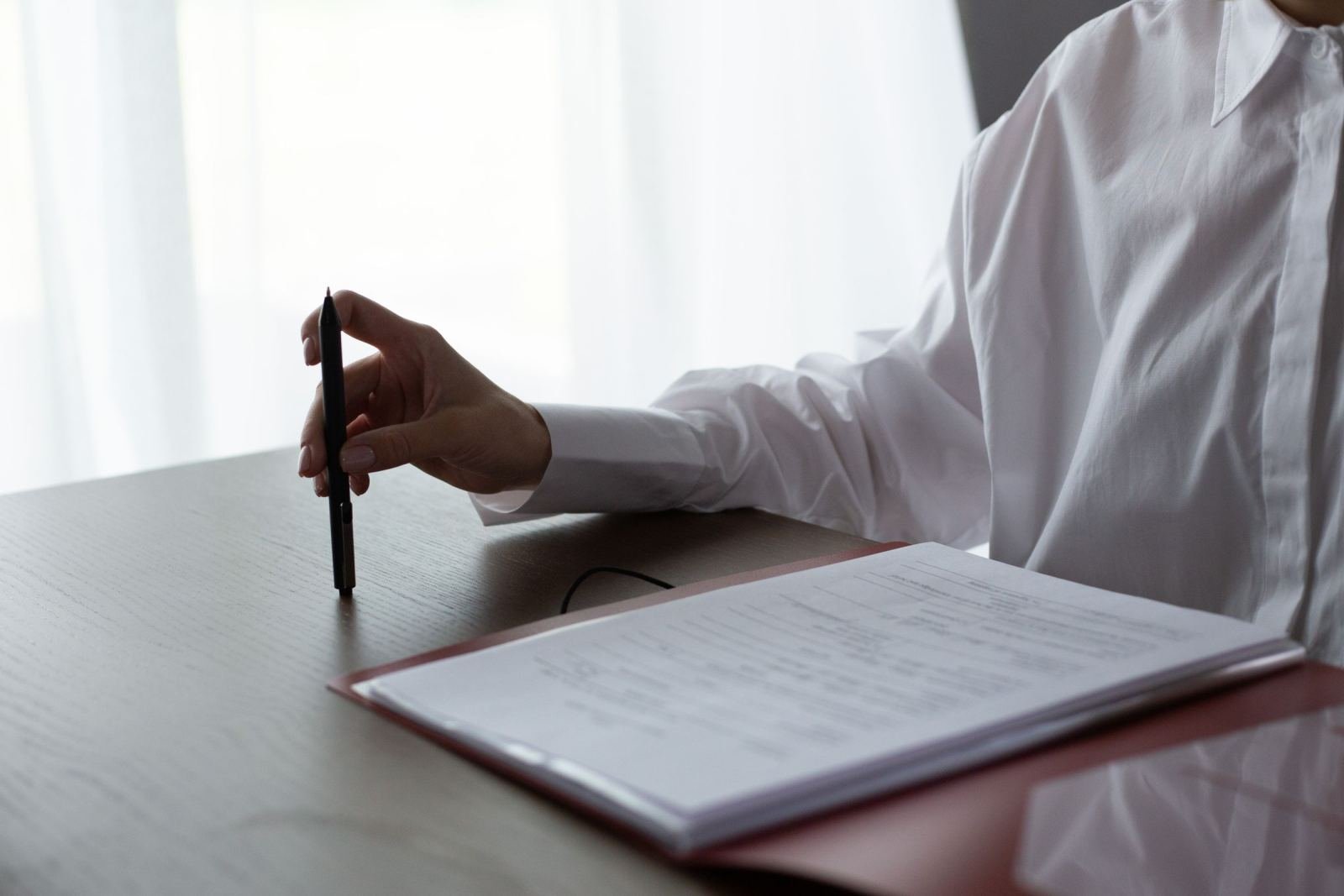 Someone in a white shirt holds a pen as they look over papers in a red folder.