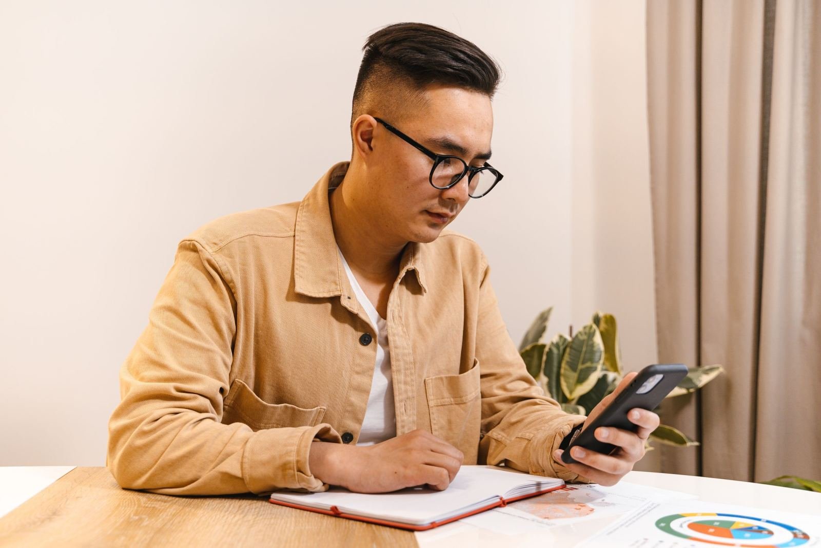 A man wearing glasses sits at a table with a notebook. He is looking at his phone.