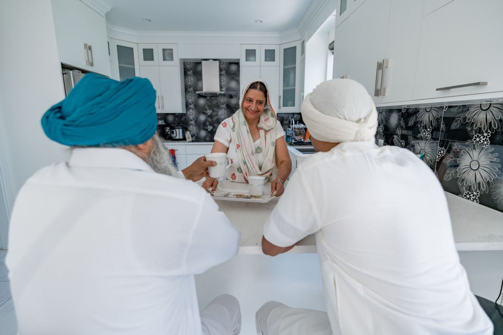 Two men in turbans sit at a kitchen counter facing a smiling woman with a tray of mugs