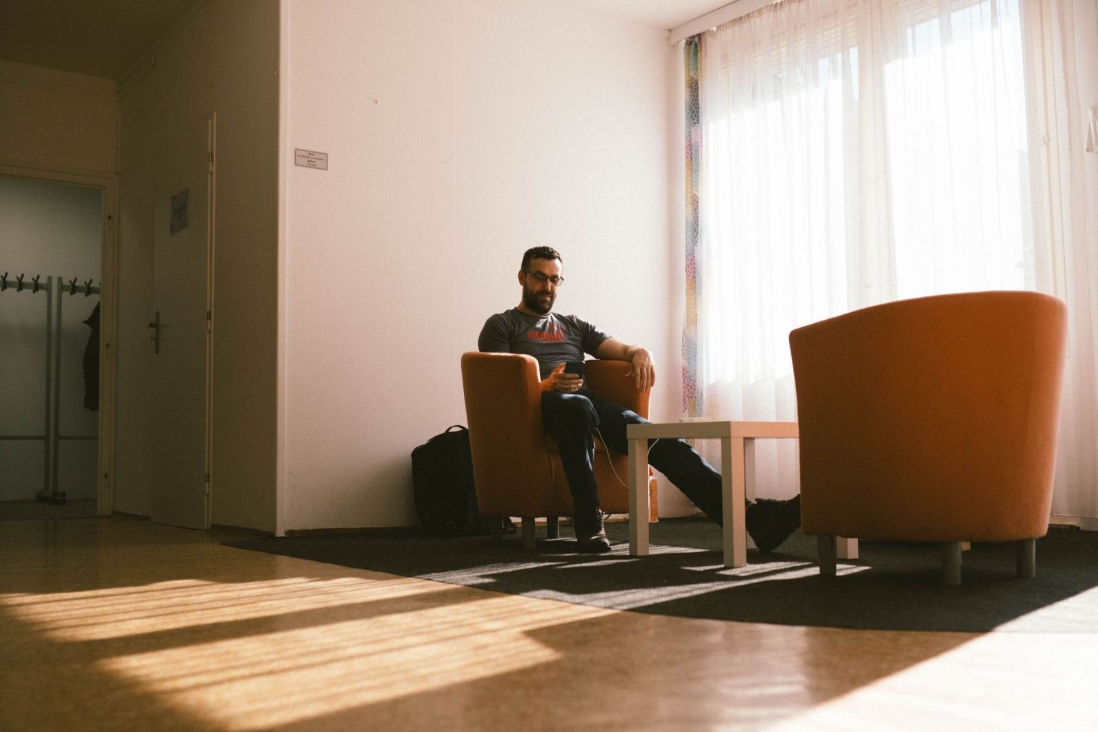 A man sits in an orange armchair in a bright room while looking at his phone